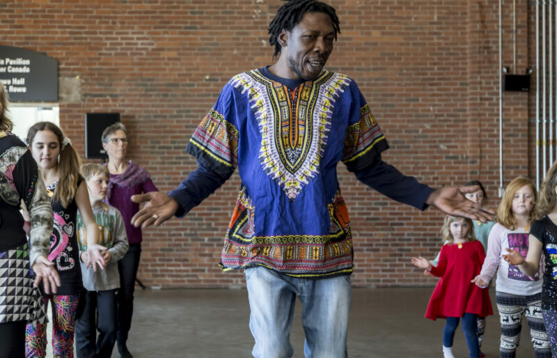 Des enfants de tous âges dansent, alignés en deux rangées aux côtés d’un danseur, tout en suivant attentivement les instructions.