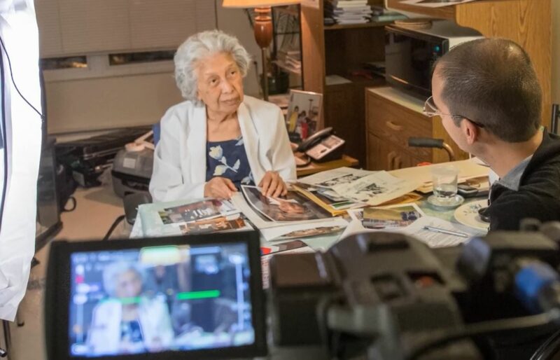 Une femme est debout à un bureau avec des photos rassemblées devant elle et elle parle à un historien oral du Musée qui lui fait face. La conversation est enregistrée avec une caméra.