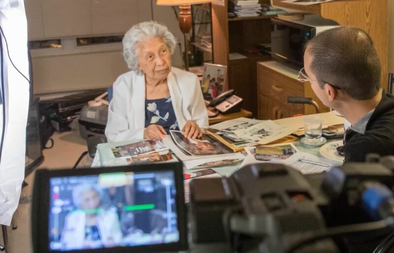 Une femme est debout à un bureau avec des photos rassemblées devant elle et elle parle à un historien oral du Musée qui lui fait face. La conversation est enregistrée avec une caméra.