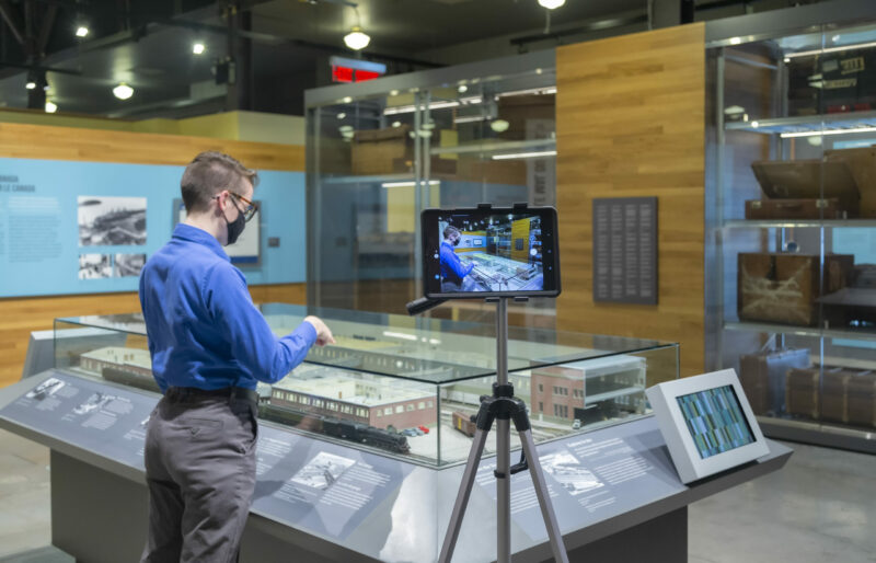 Un interprète du patrimoine se tient devant la maquette du hangar d’immigration du Quai 21 tandis qu’un iPad enregistre sa présentation.