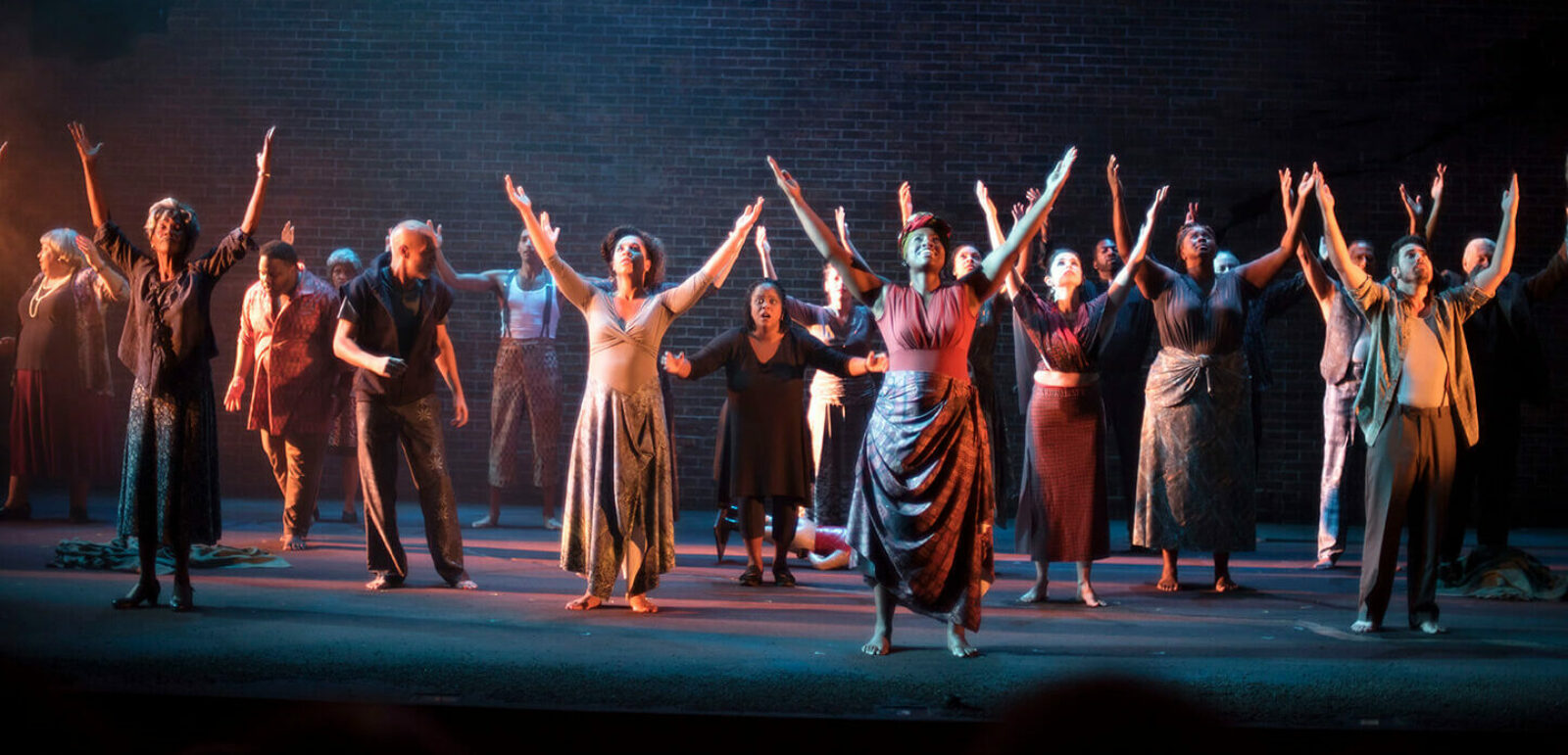 Two dozen actors on an empty stage with their hands raised towards the sky.