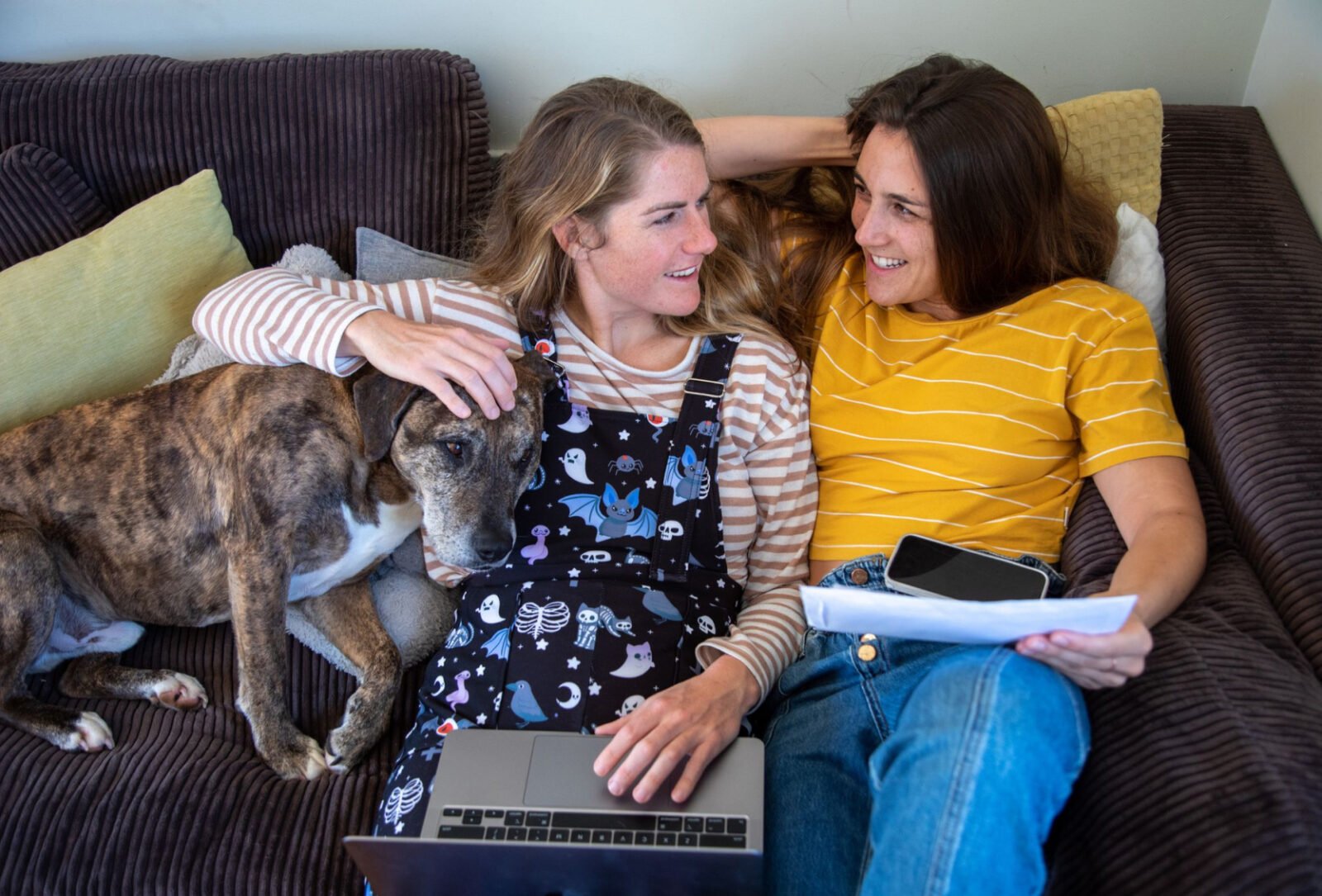 Two women and a dog sitting on their couch with a laptop